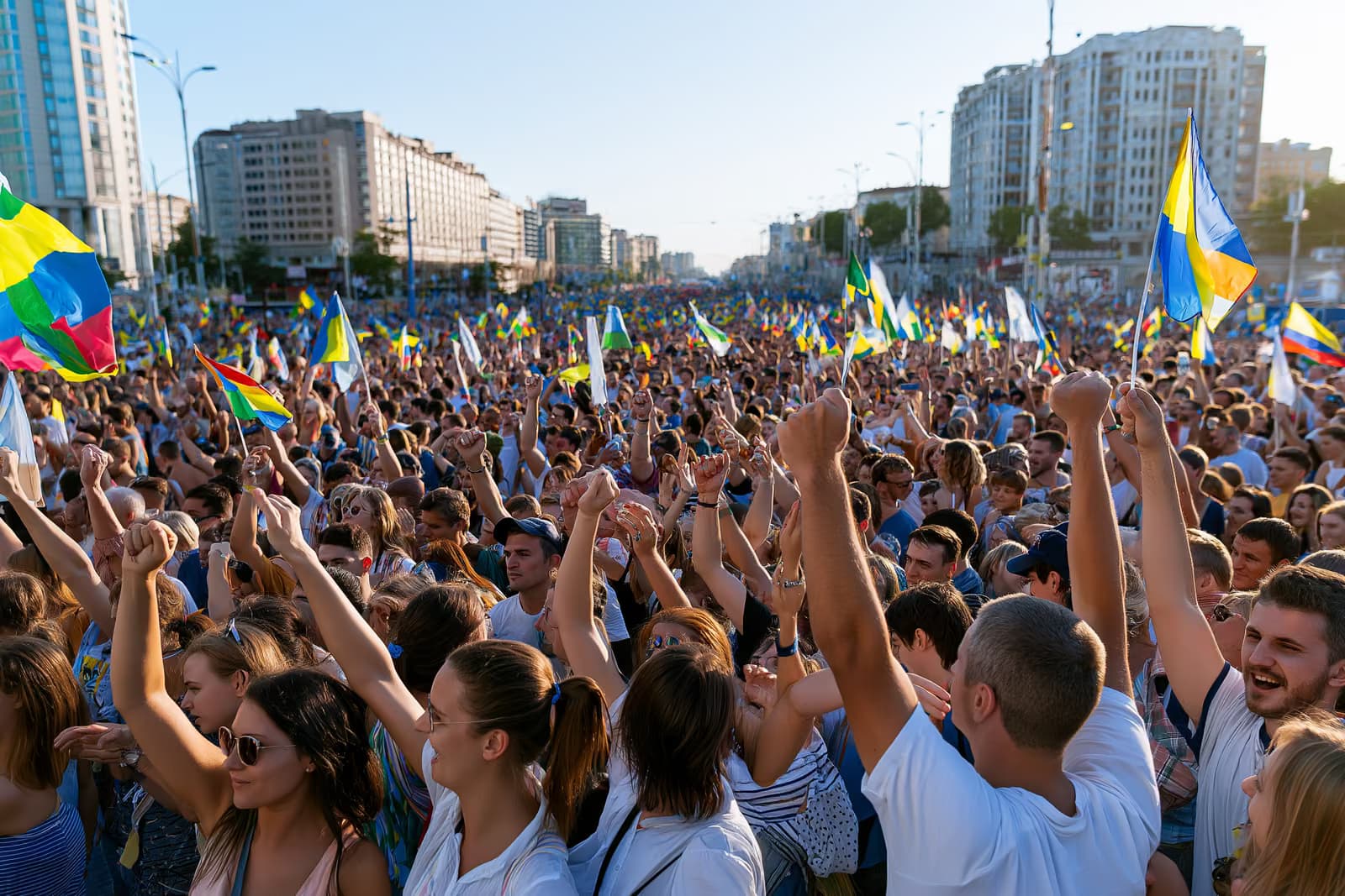 Une foule immense remplit une rue de la ville sous un ciel d'été radieux, agitant des drapeaux colorés.