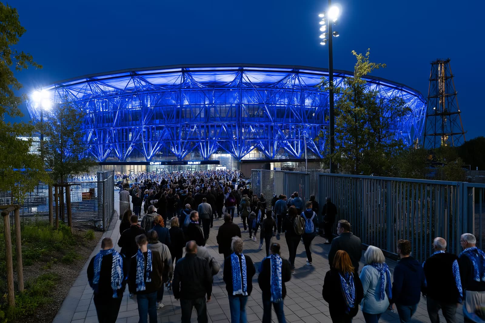 Un stade de football de la Ruhr s'illumine d'un bleu royal la nuit, tandis que des supporters en écharpes se dirigent vers les portes après le transfert de Gosens à Schalke.